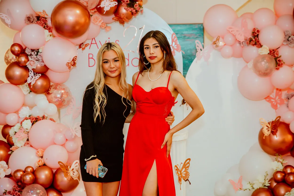 Two friends pose together against pink and rose gold balloon backdrop at celebration.