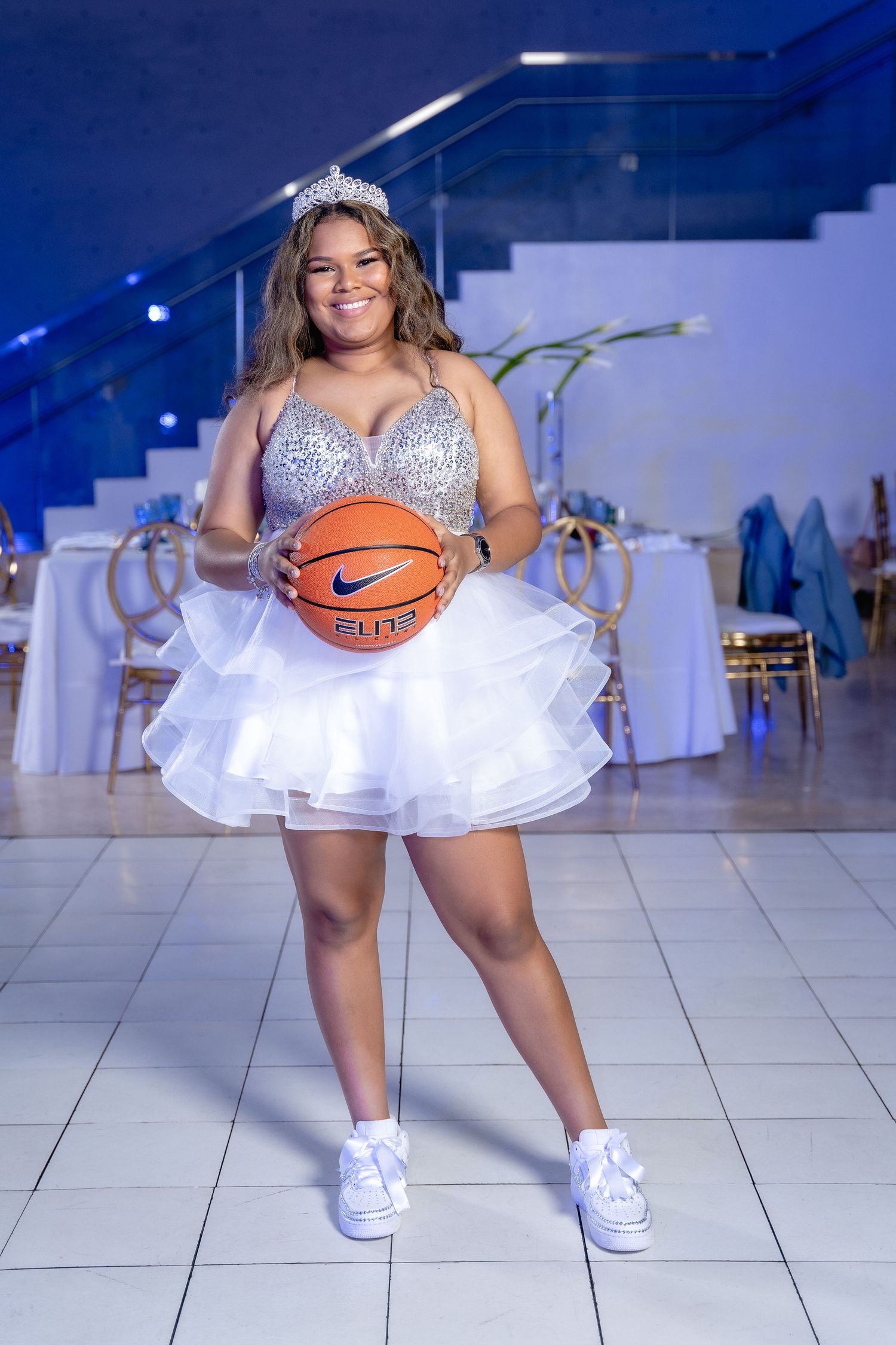 Someone poses in a sparkly white dress and sneakers while holding a basketball in an elegant event space with blue lighting.