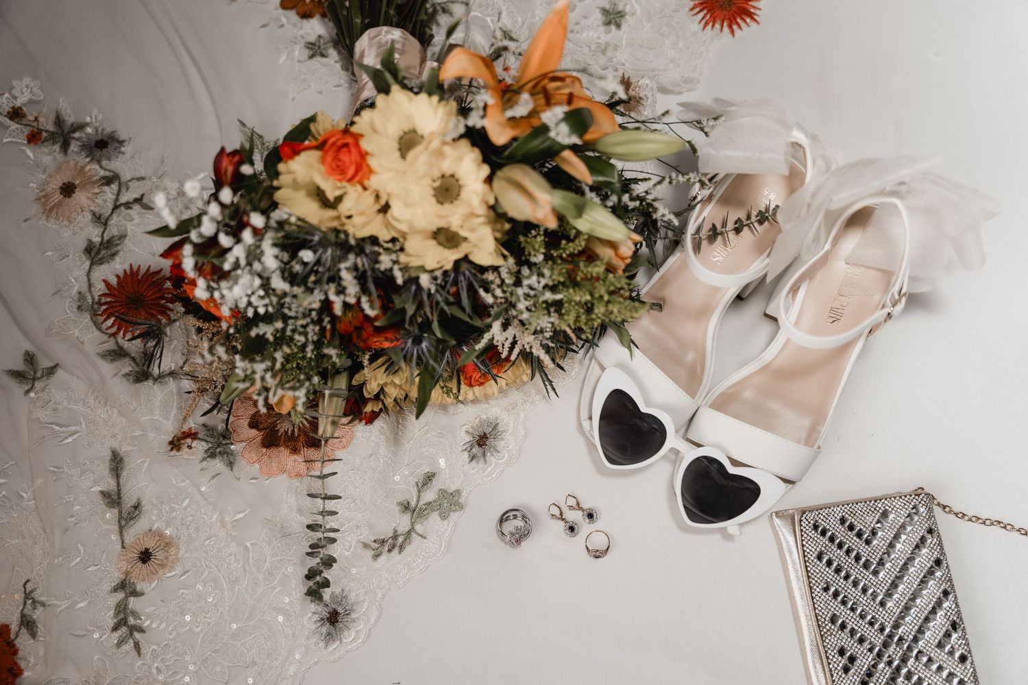 Wedding flatlay with autumn-toned floral bouquet, white bridal shoes, sunglasses and clutch on textured background.