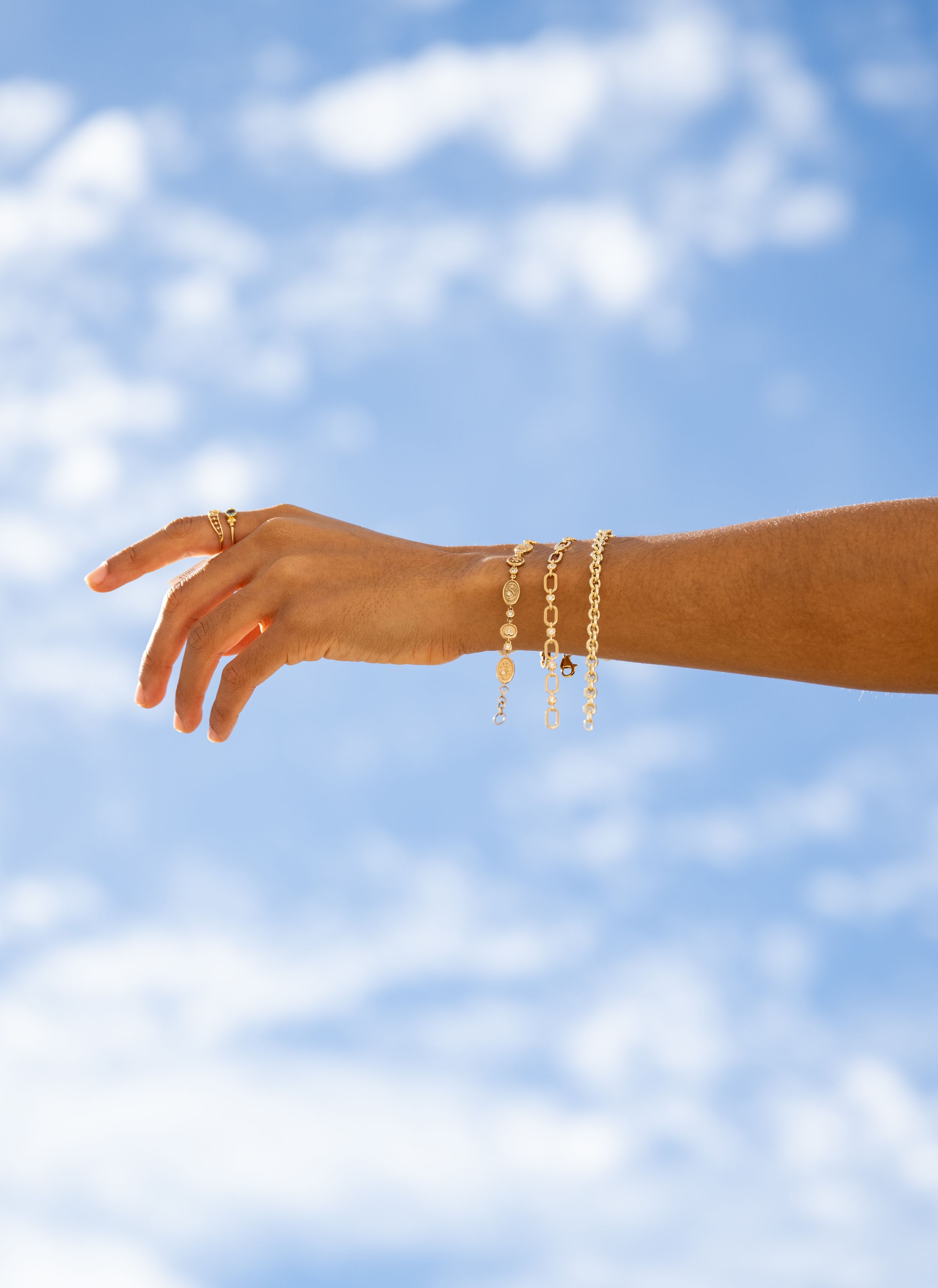 An elegant hand wearing delicate jewelry bracelets extends gracefully against a blue sky with white clouds.