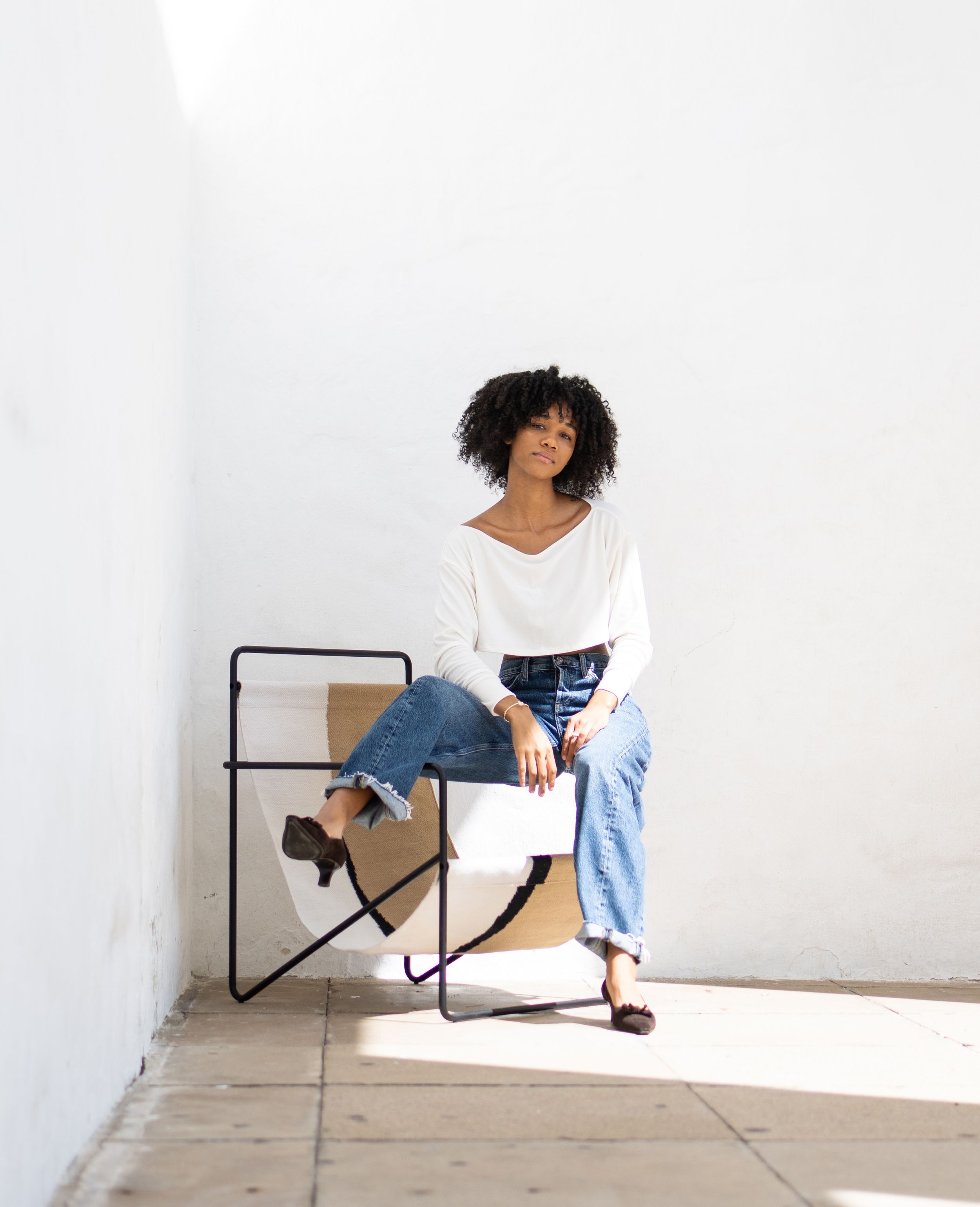 Minimalist portrait in white v-neck sweater and jeans, seated on modern chair in sunlit space.