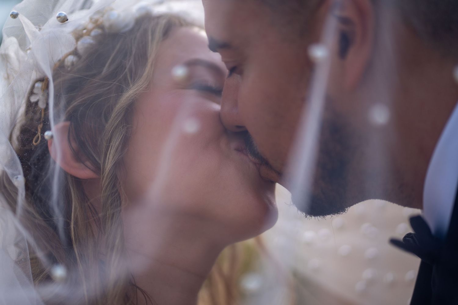 Close-up of a bride and groom kissing under a pearl-detailed veil. Romantic wedding photography capturing intimate and timeless love.