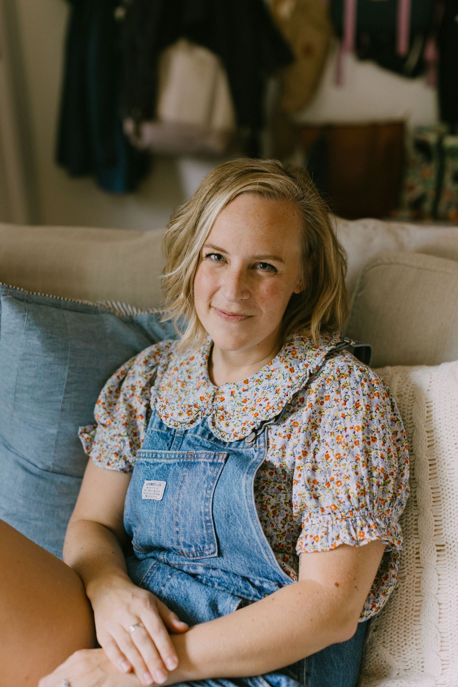 A blonde woman wearing denim overalls and a floral print top sits casually on a couch in a warm indoor setting.