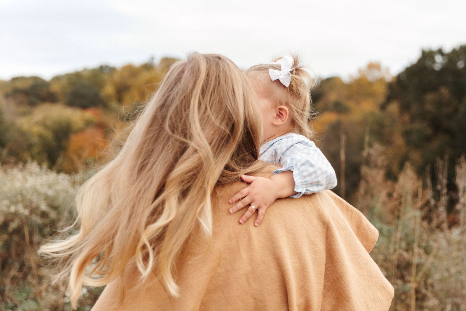 A tender autumn moment captured from behind showing a parent holding a small child outdoors.