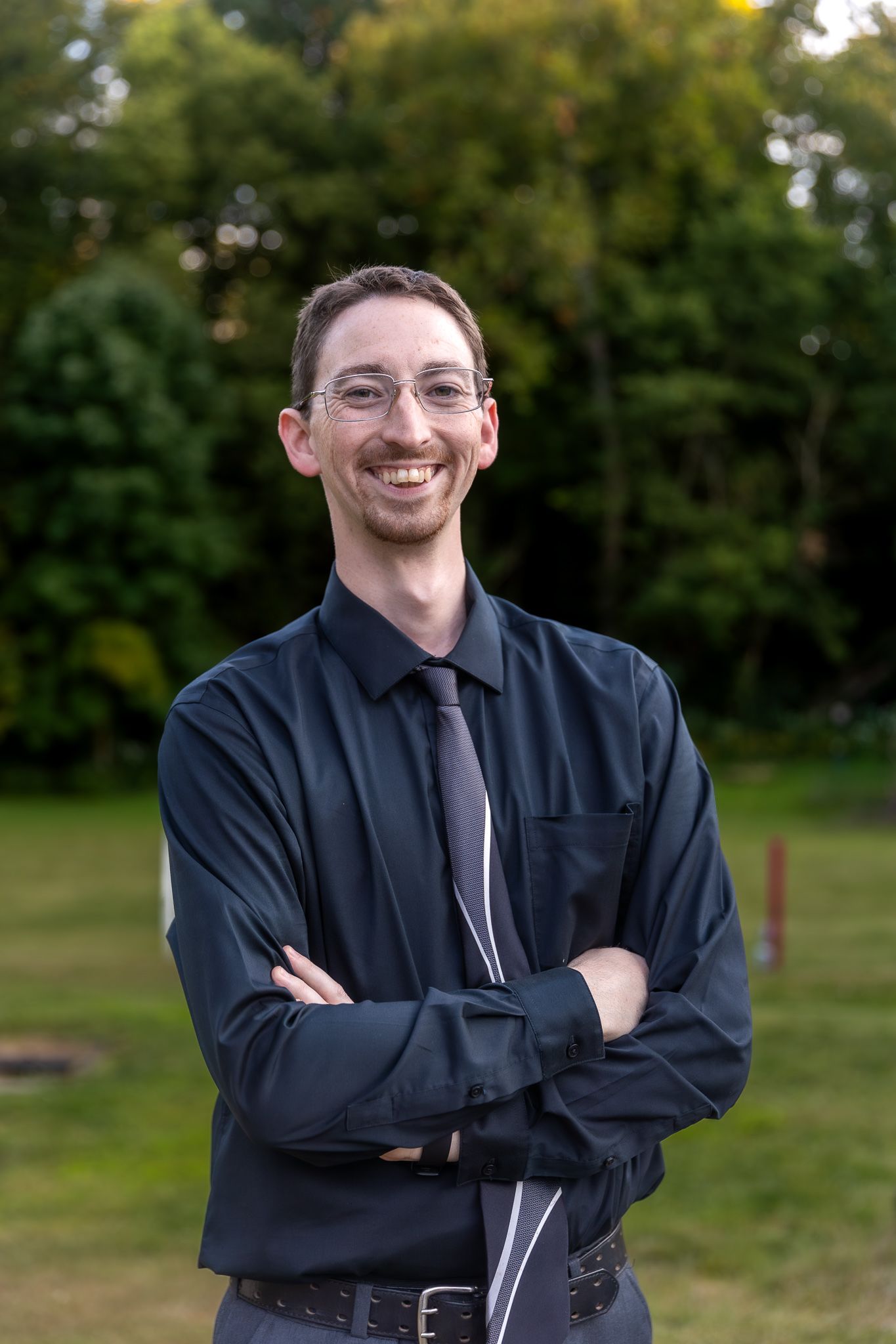 A person in business attire stands with arms crossed outdoors with trees in background.