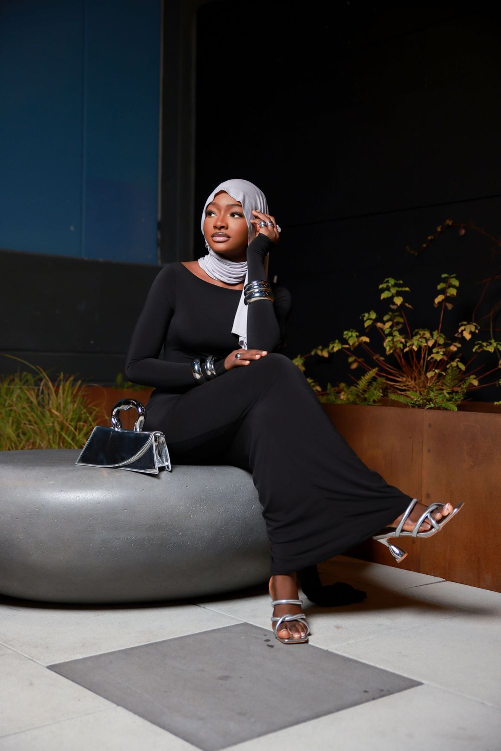A fashion model in a black outfit and white headscarf poses on a concrete bench with a small handbag.