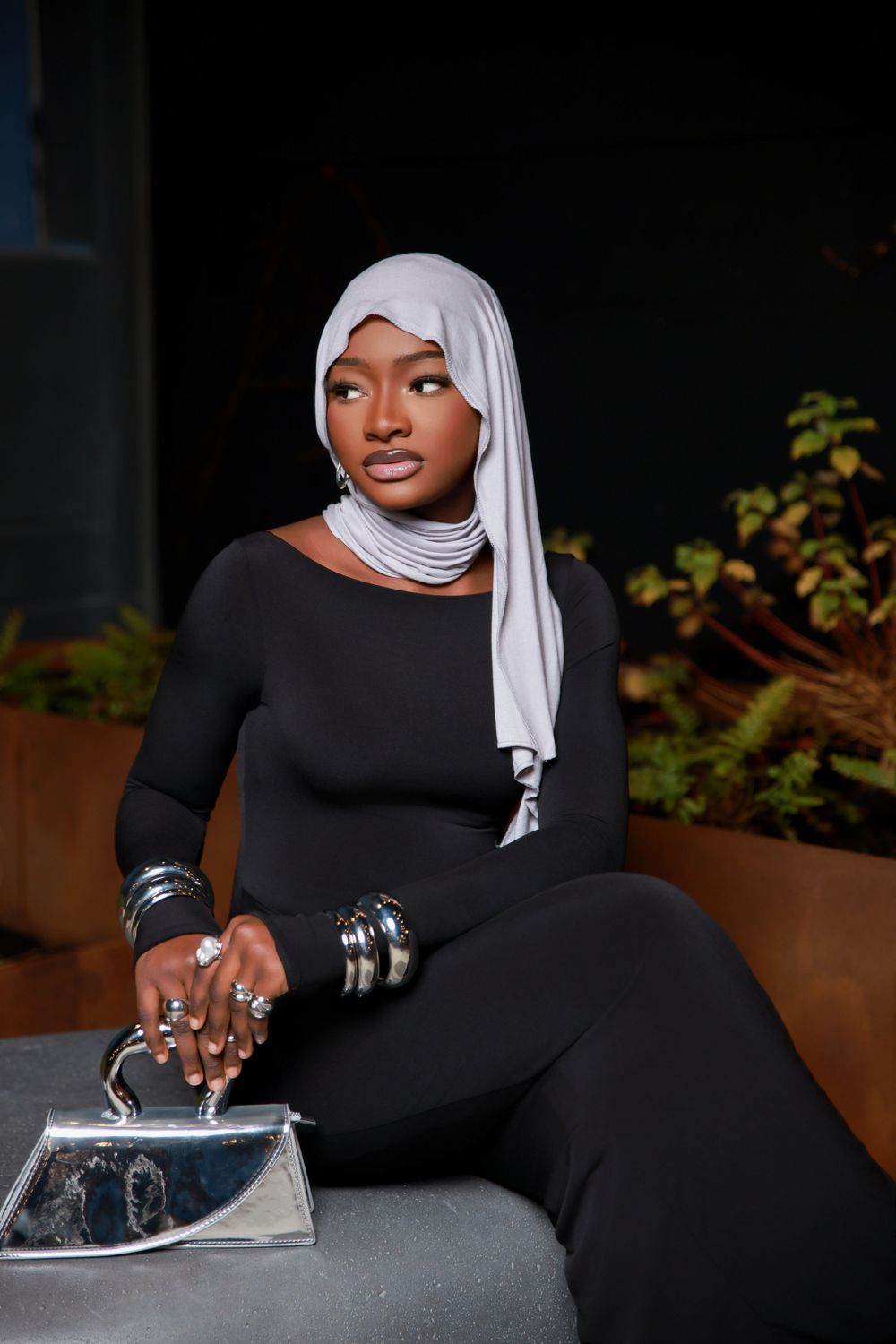 A fashionable pose on a concrete bench wearing a black ensemble with white headscarf and silver accessories.