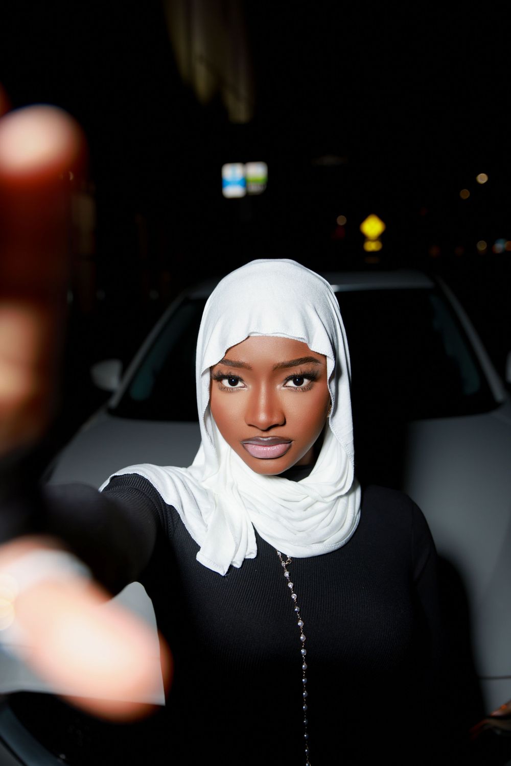Nighttime portrait wearing white hijab and black outfit against car background.