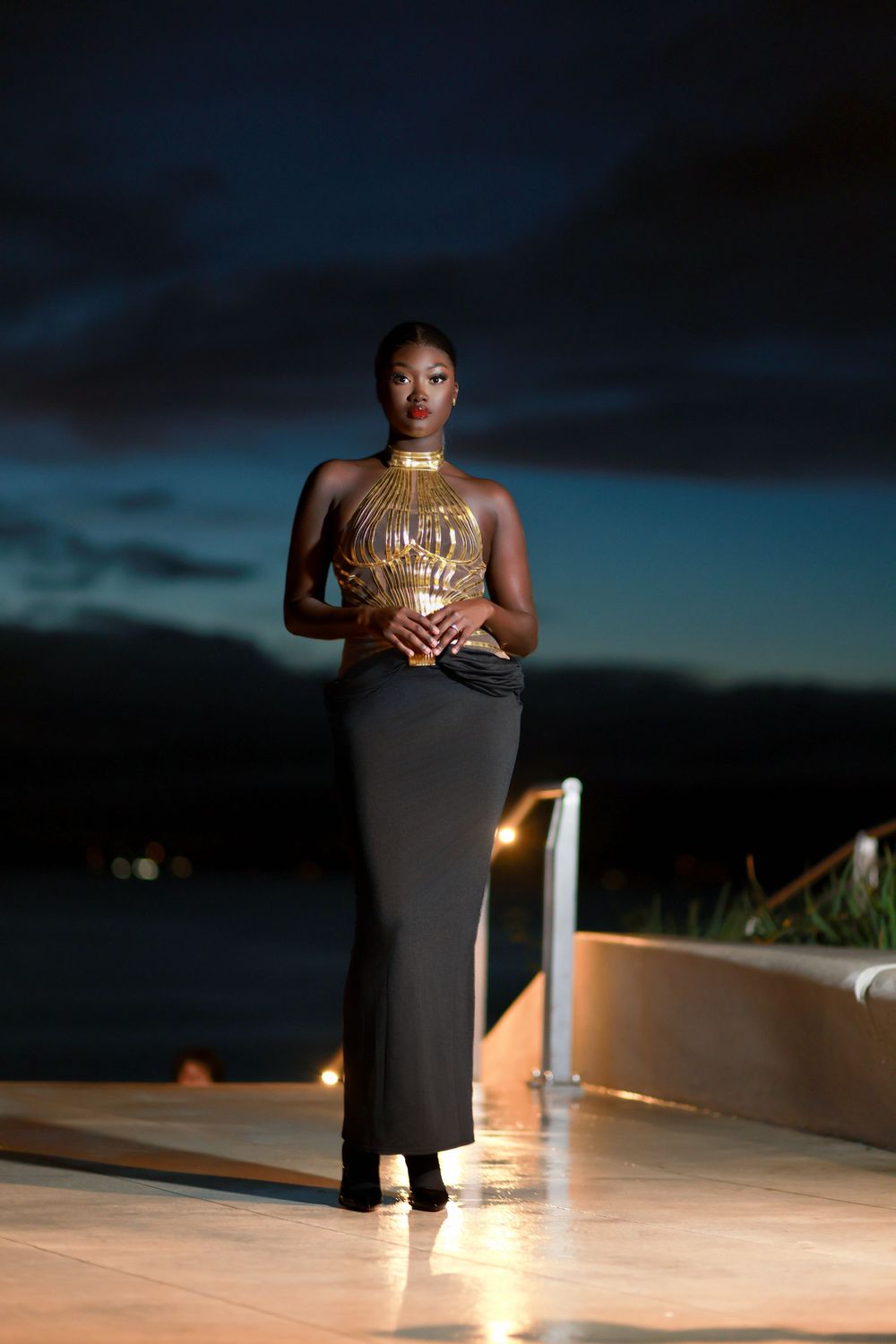Elegant evening pose in black dress with gold halter top against sunset sky.