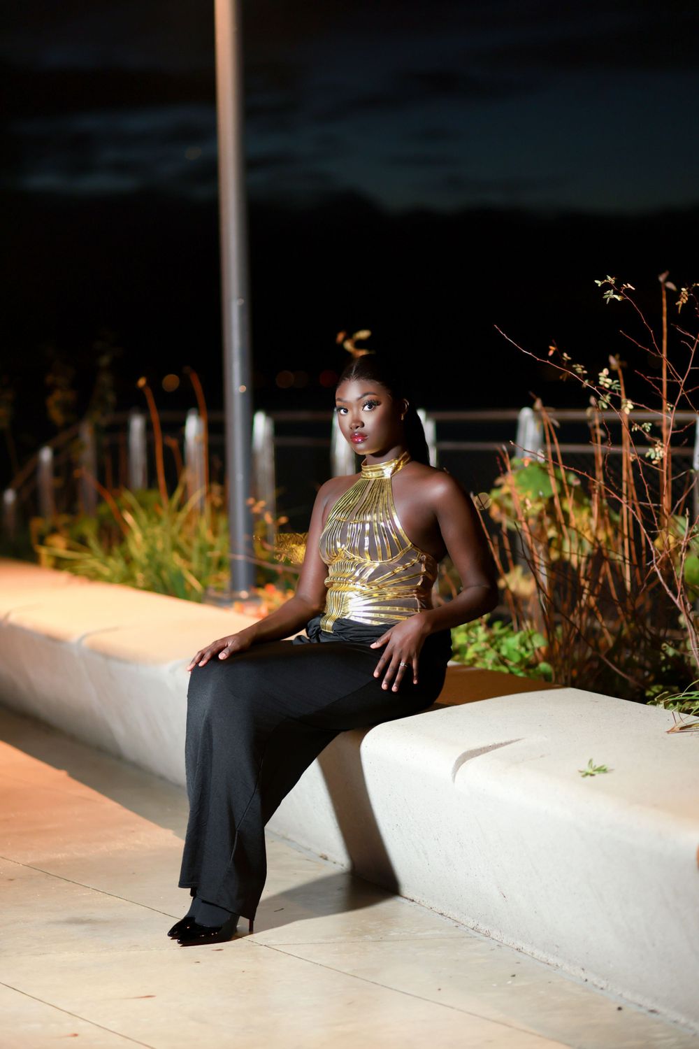 Outdoor evening portrait on white ledge wearing metallic halter top and black pants.