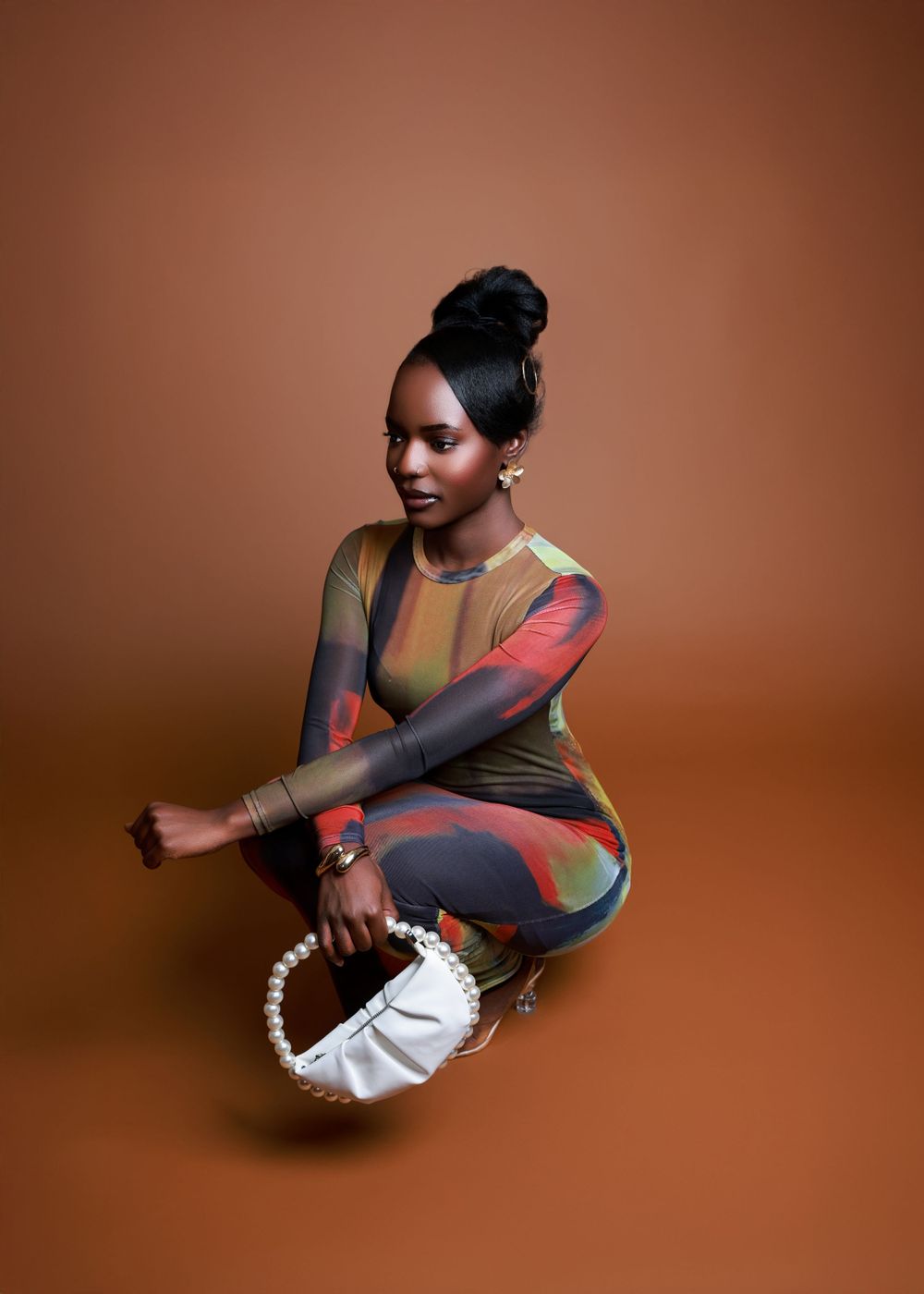 A fashion portrait featuring a colorful patterned bodysuit and white pearl-handled handbag against brown backdrop.