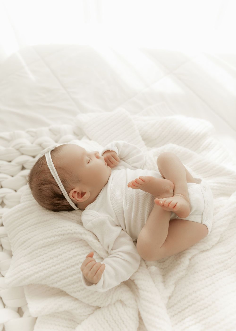 A sleeping baby in white clothing lies peacefully on soft white bedding in a series of connected photos.