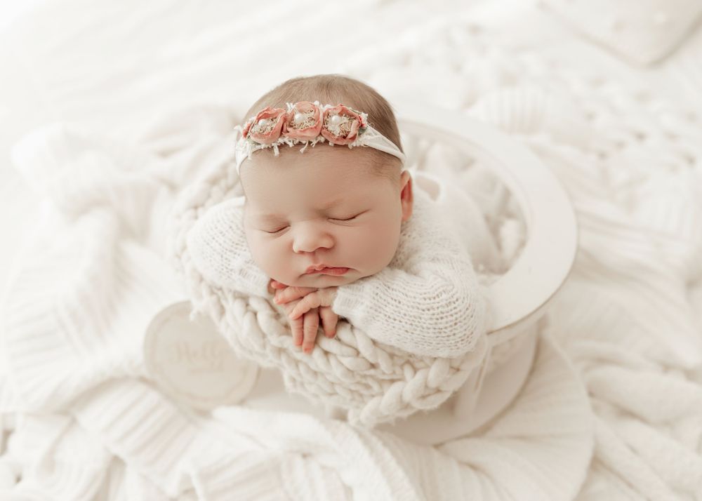 Sleeping newborn wrapped in soft white lace wearing a delicate rose gold crown headband on white textured backdrop.