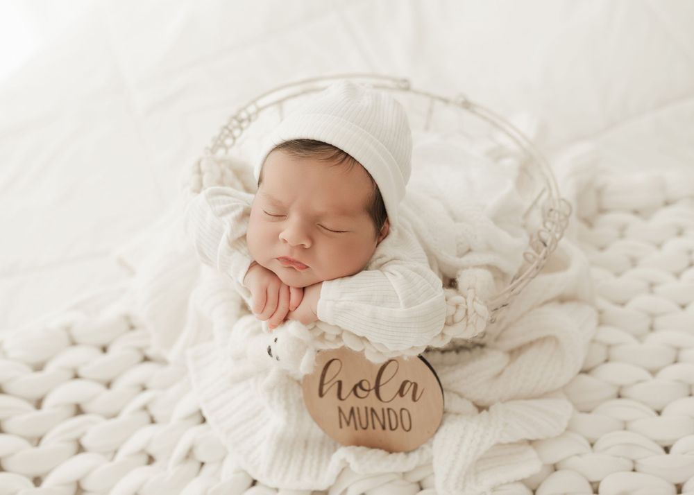 Sleeping newborn baby wrapped in soft white swaddle blanket with matching hat rests peacefully on white backdrop.