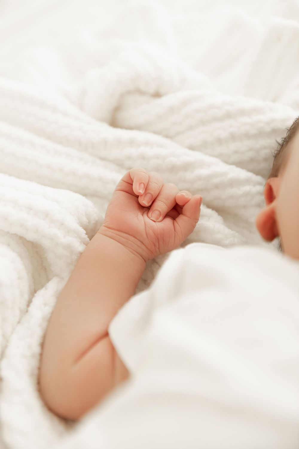 A tiny baby hand reaches out from soft white bedding in a tender moment of rest and comfort.