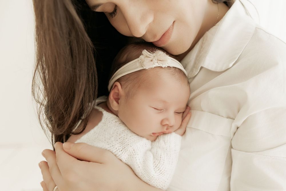 A tender moment between mother and sleeping newborn baby in white, captured in soft natural light.