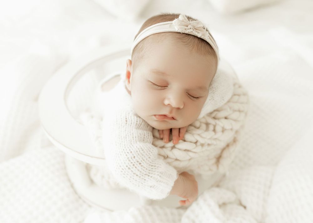 A sleeping newborn baby dressed in white rests peacefully on soft white bedding.