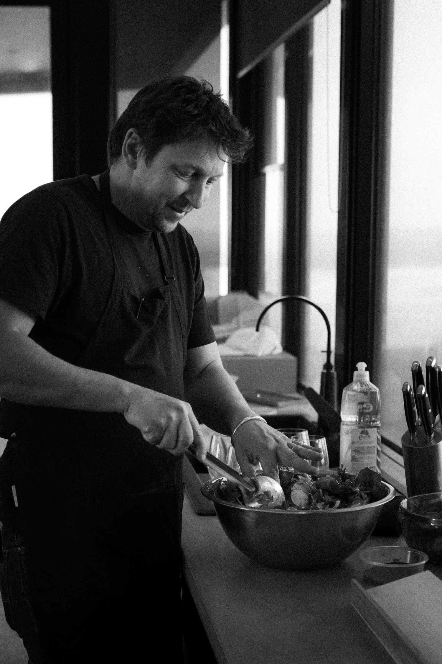 Black and white photo of a man preparing food in a bowl at a kitchen counter.