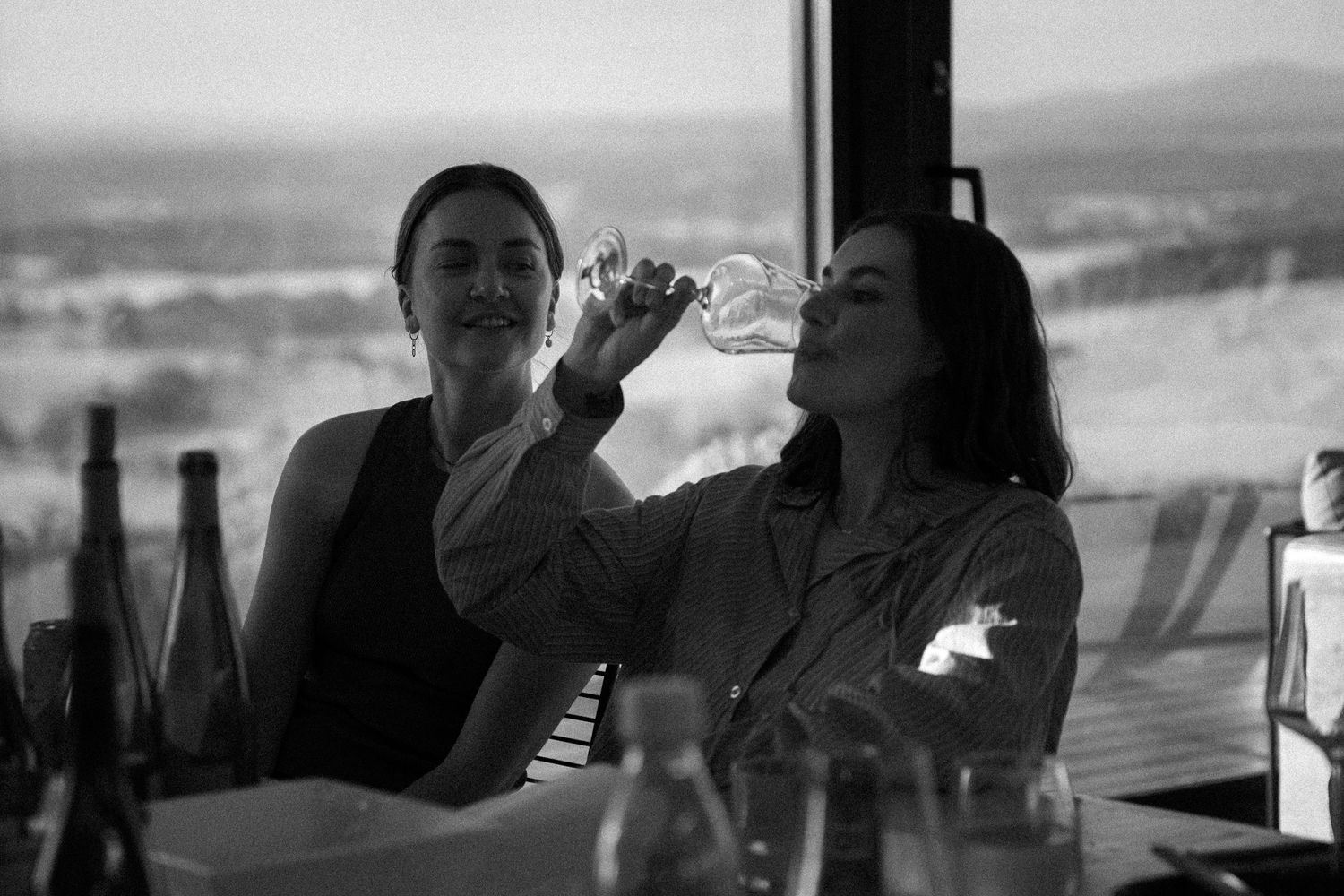 Black and white photo of two women at a table, one drinking wine with a scenic view behind.