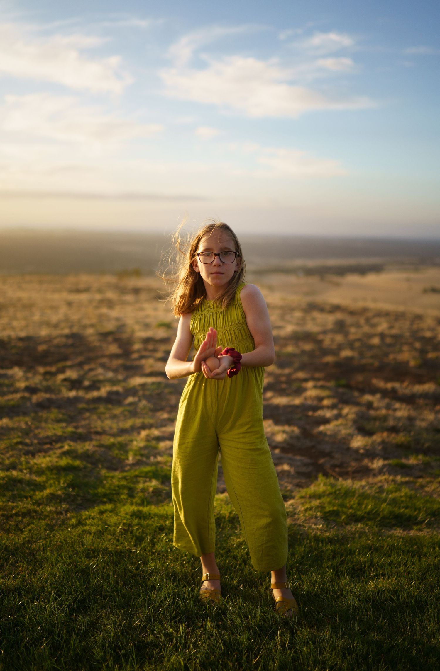 Young girl in a green jumpsuit standing in a sunlit field holding a red apple.