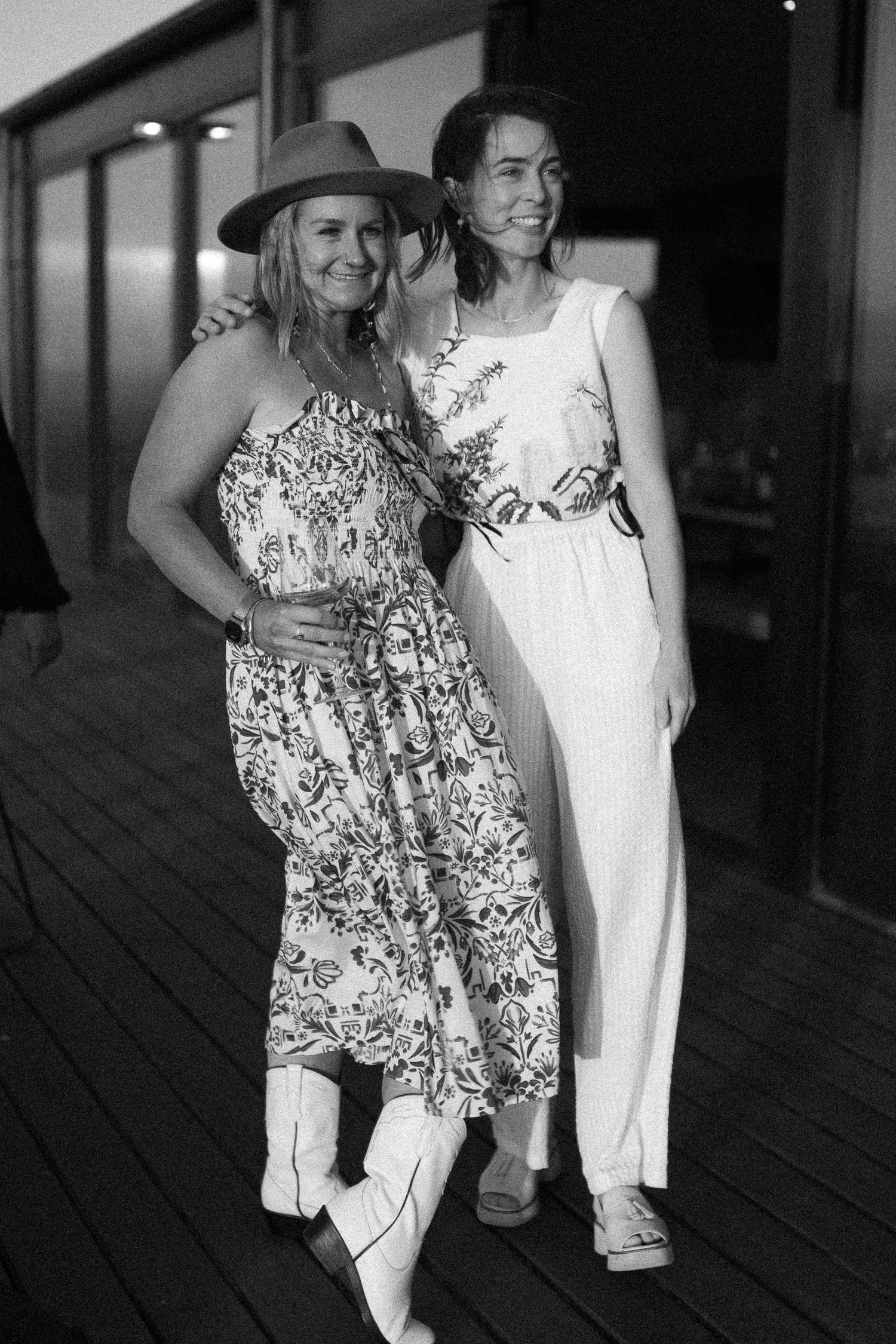 Black and white photo of two women posing together on a deck, one wearing a hat and floral dress.