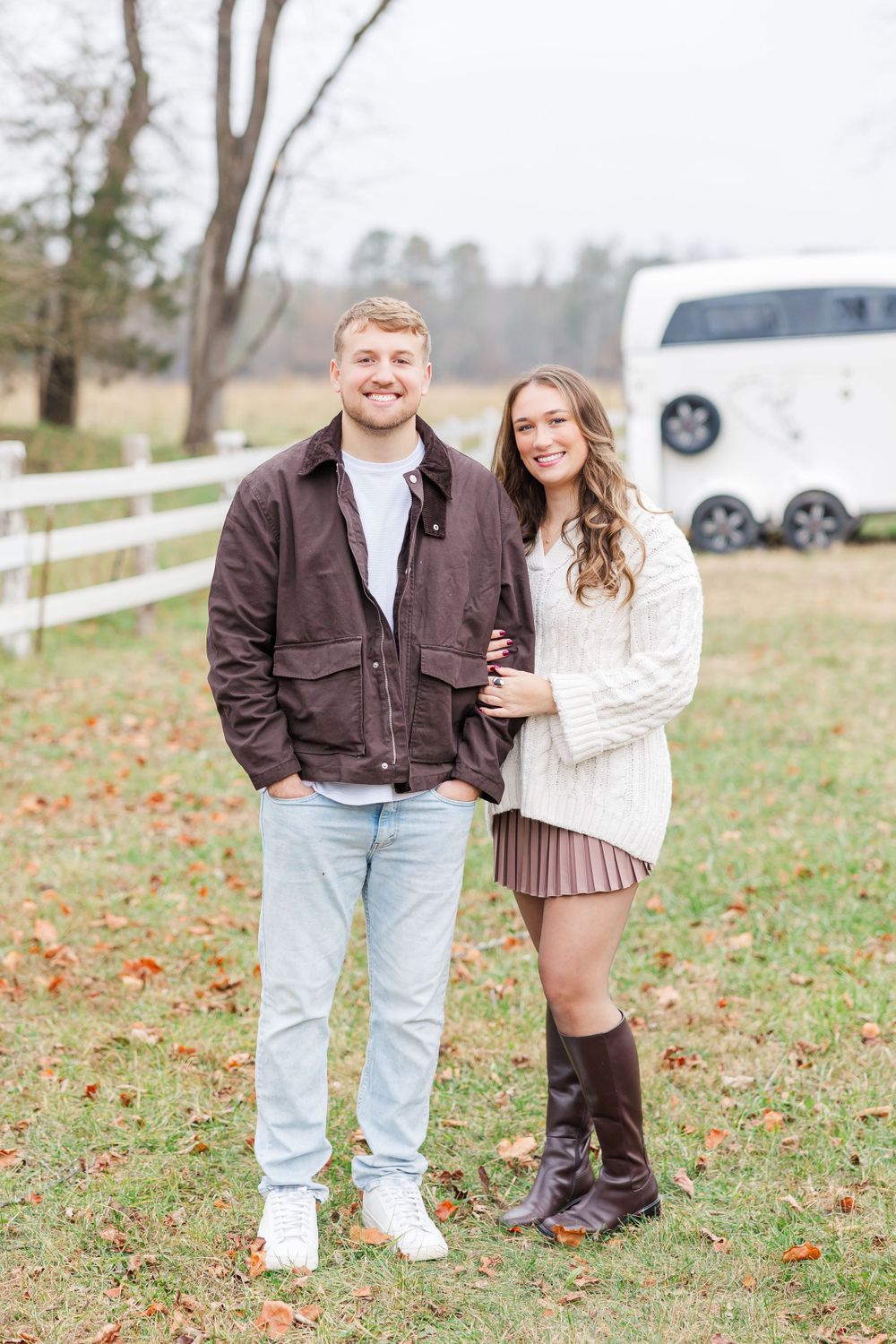 Couple in casual fall attire standing together in front of white fence on autumn day.
