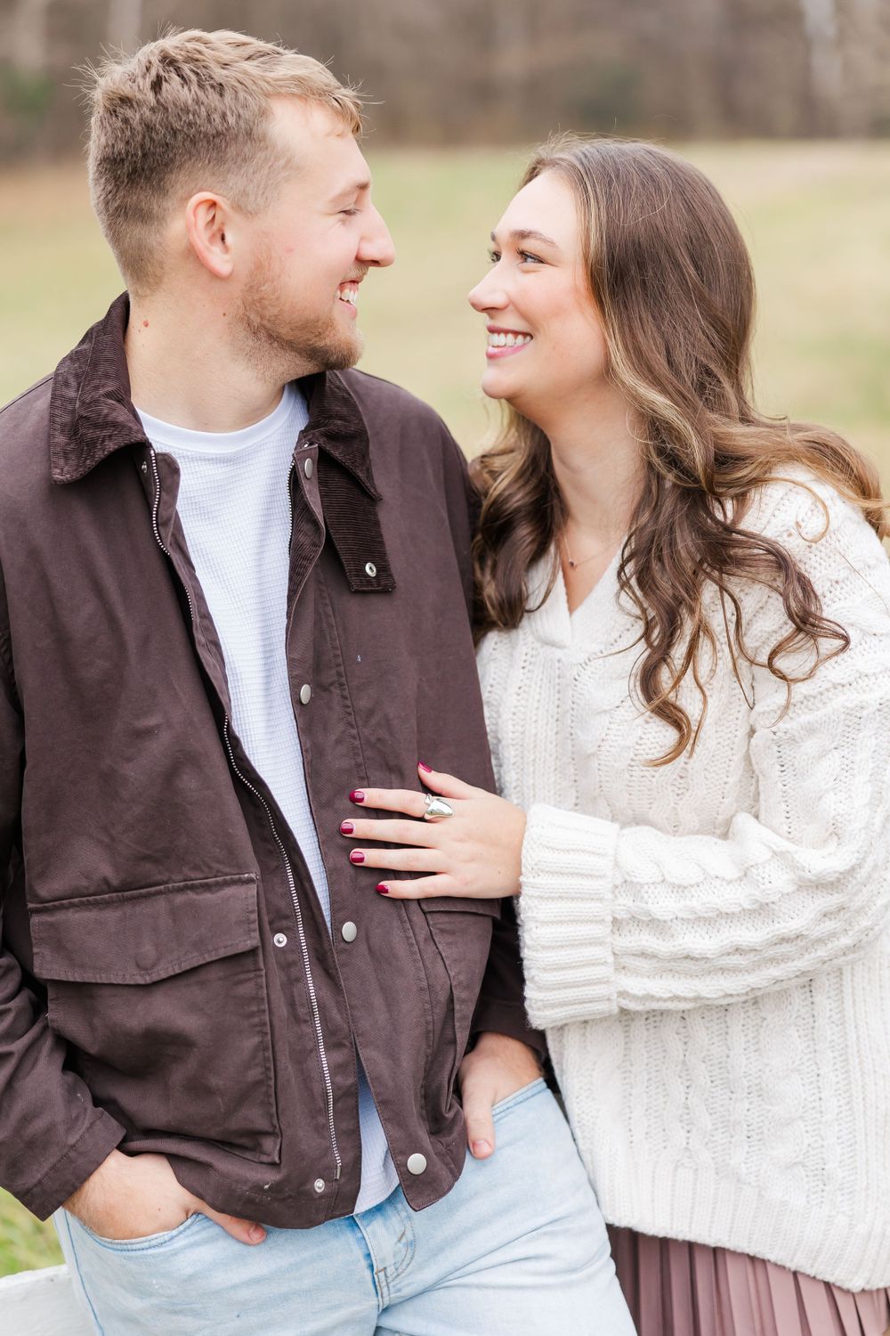 Couple in casual winter outfits smiling at each other against rural backdrop.