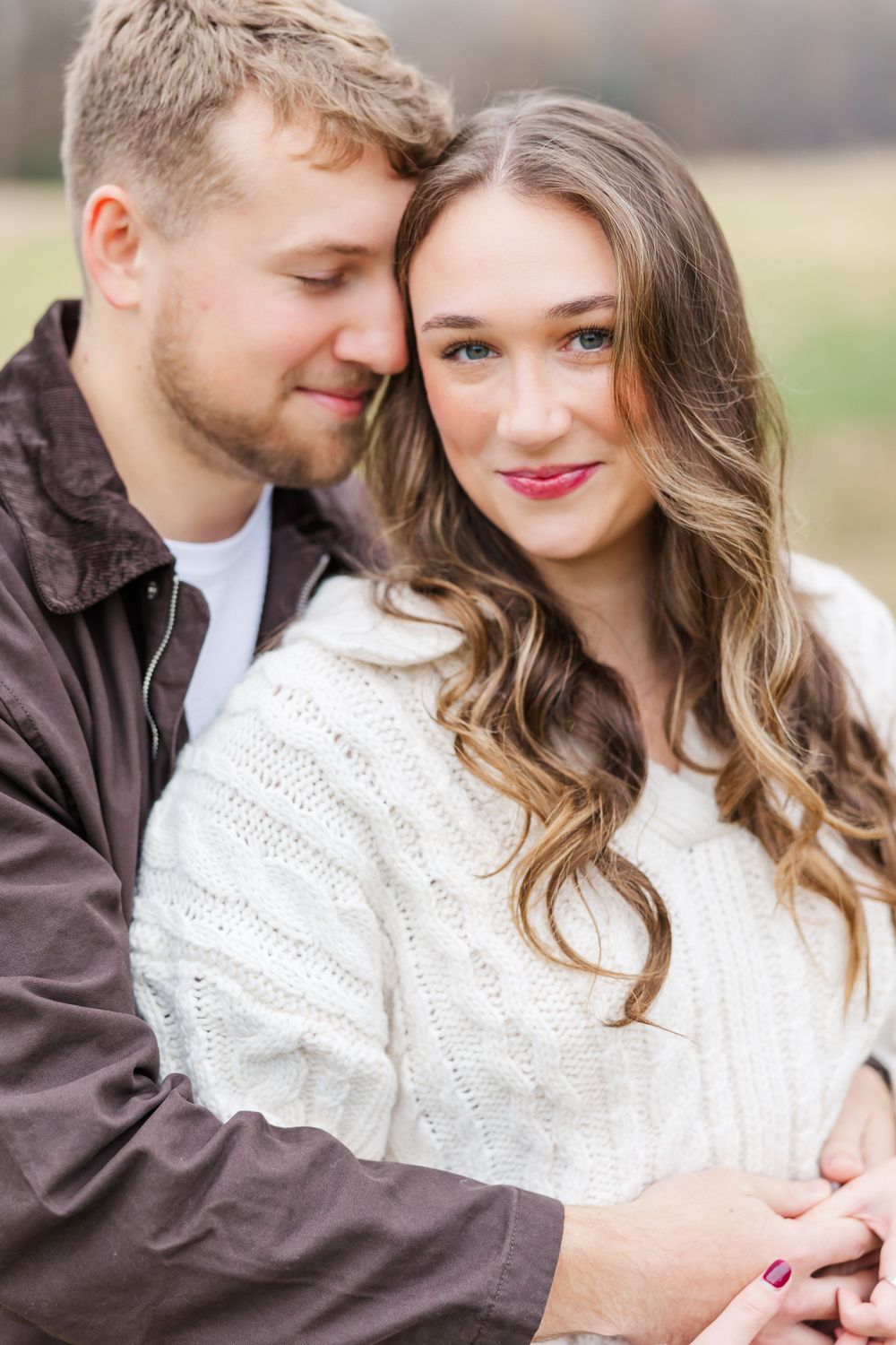 A romantic couple embraces outdoors, with her wearing a white sweater and him in a brown jacket.