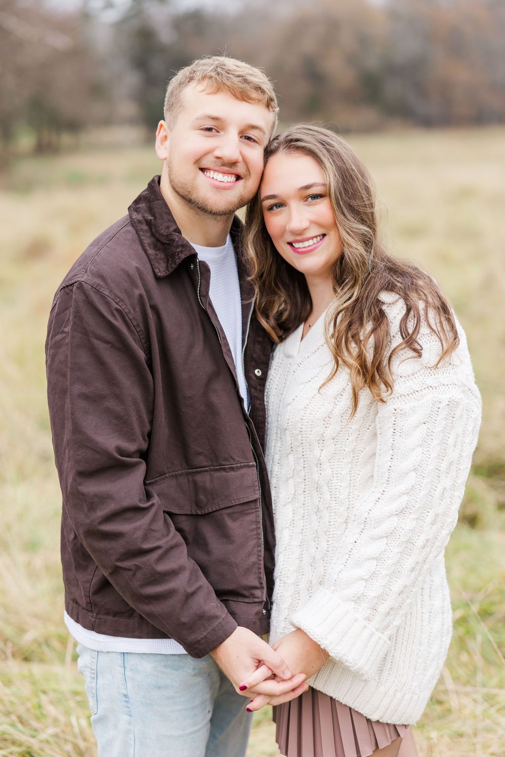 A happy couple smiles at the camera while embracing in a field wearing casual fall attire.