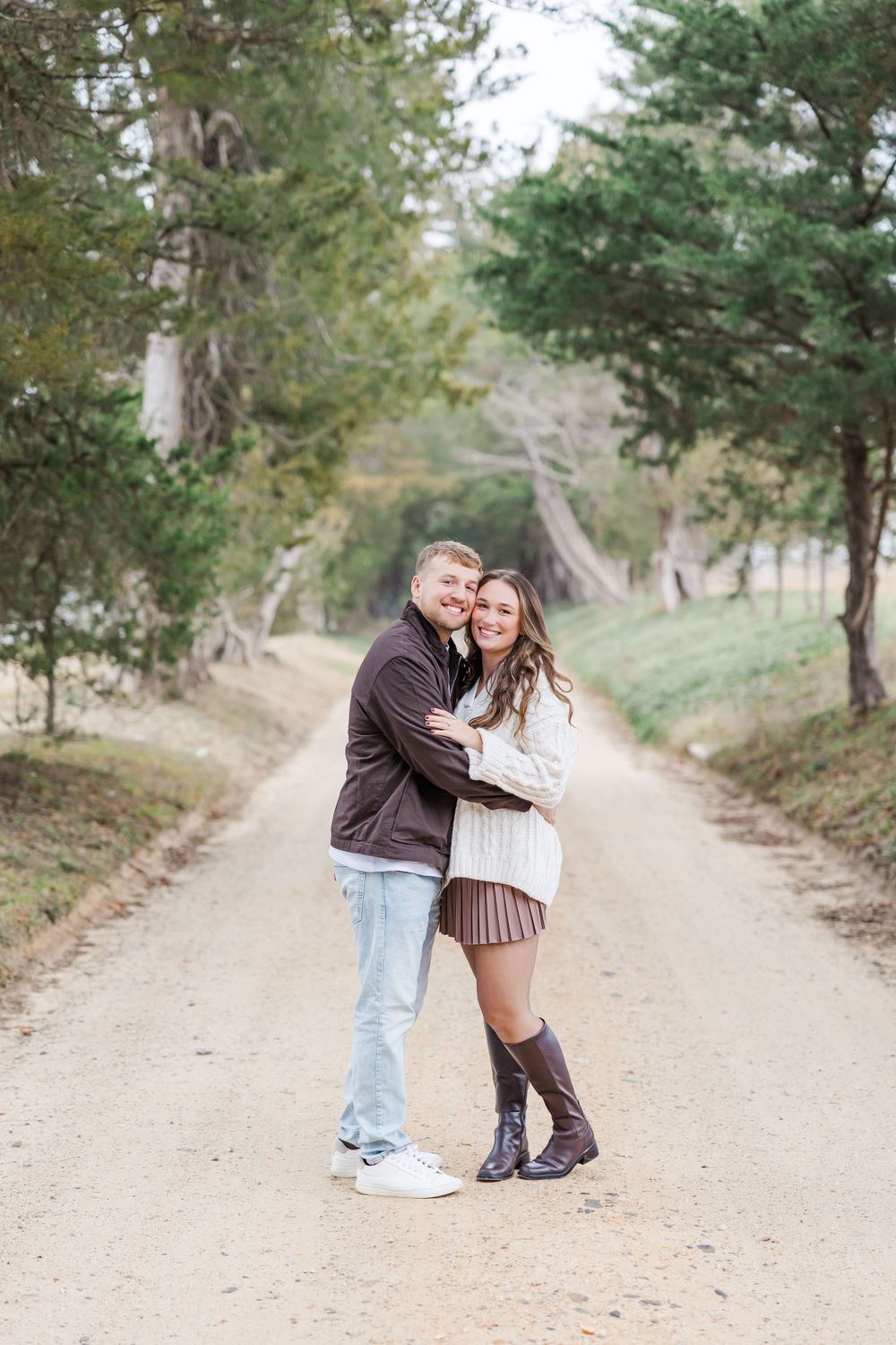 A couple poses together on a dirt path surrounded by pine trees.