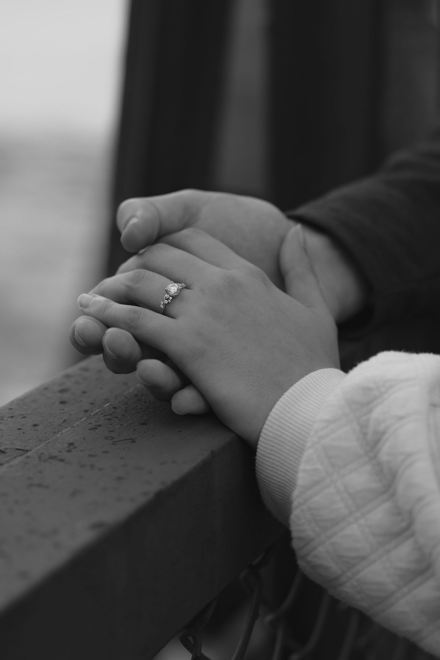 Black and white photo of two hands holding together on a railing, with a diamond engagement ring visible.
