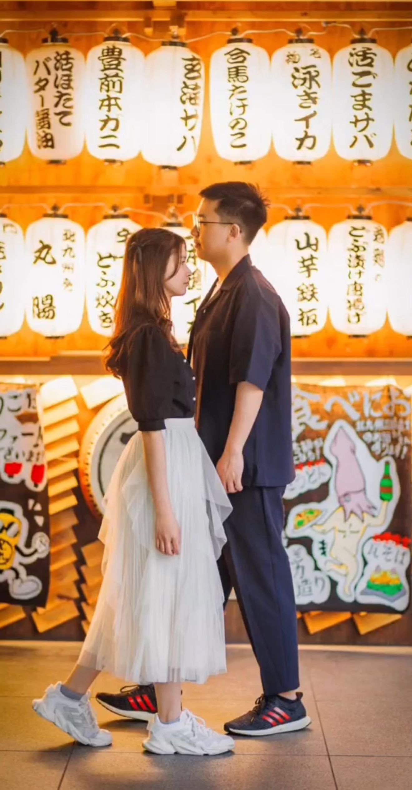 A couple poses in front of traditional Japanese paper lanterns wearing casual modern clothing and sneakers.
