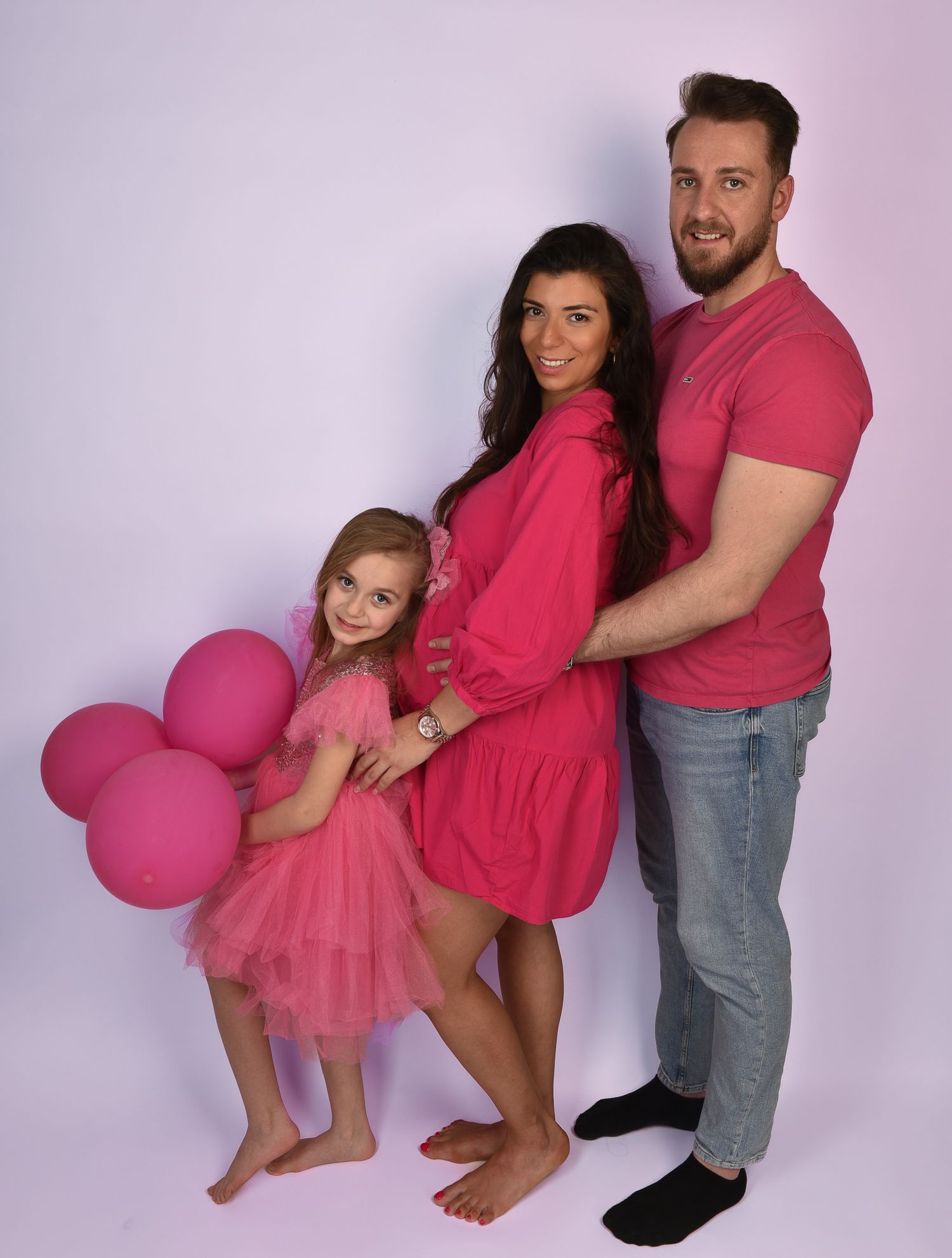 Familienfotografie, Family wearing matching pink outfits poses together with balloons against purple backdrop.