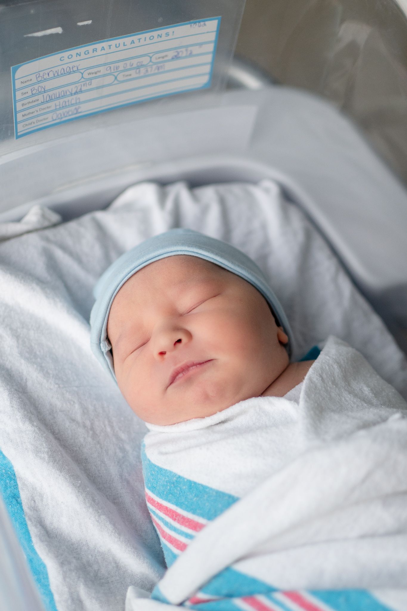 A sleeping newborn infant wearing a light blue cap rests peacefully swaddled in white and blue hospital blankets.