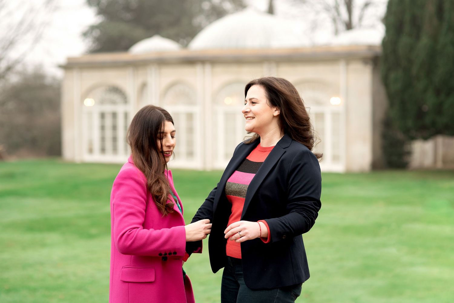 Two figures in conversation standing outdoors on a green lawn in front of a stone gazebo, one wearing pink and one in navy.