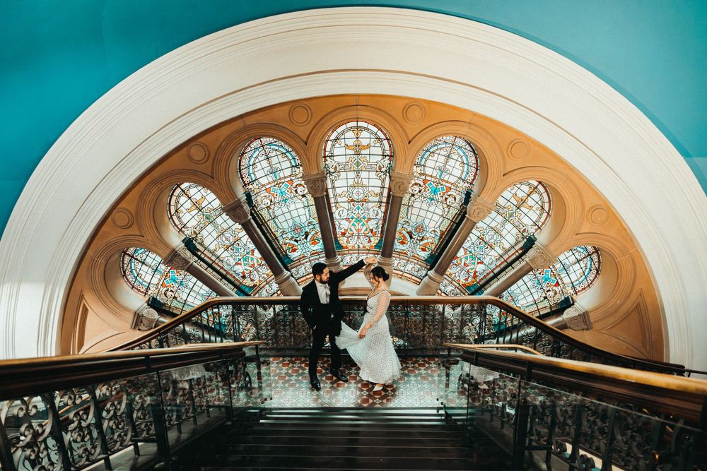 Artistic overhead shot of a couple on vintage staircase with fan-shaped stained glass window above.