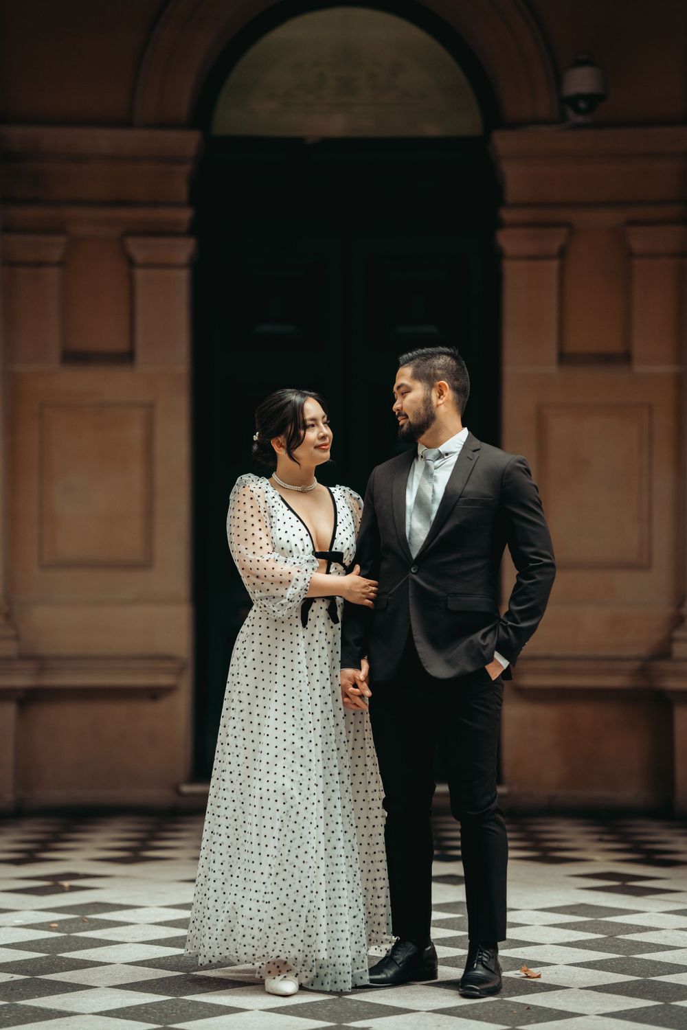 A couple shares an intimate moment on a checkered floor in front of an elegant arched doorway.