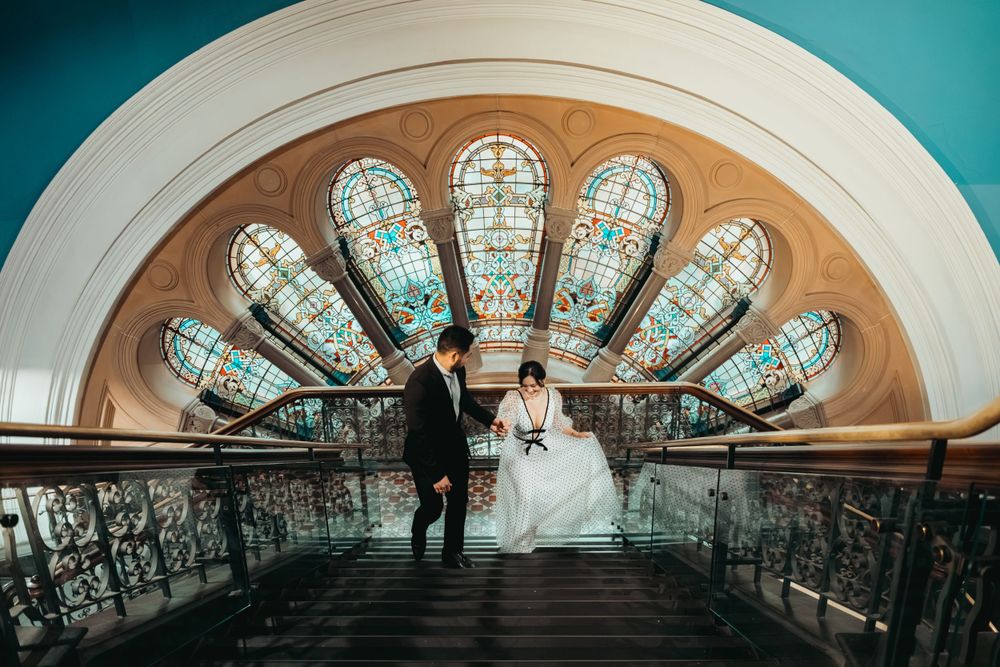 A couple poses against a stunning art nouveau stained glass fan window in a historic building.