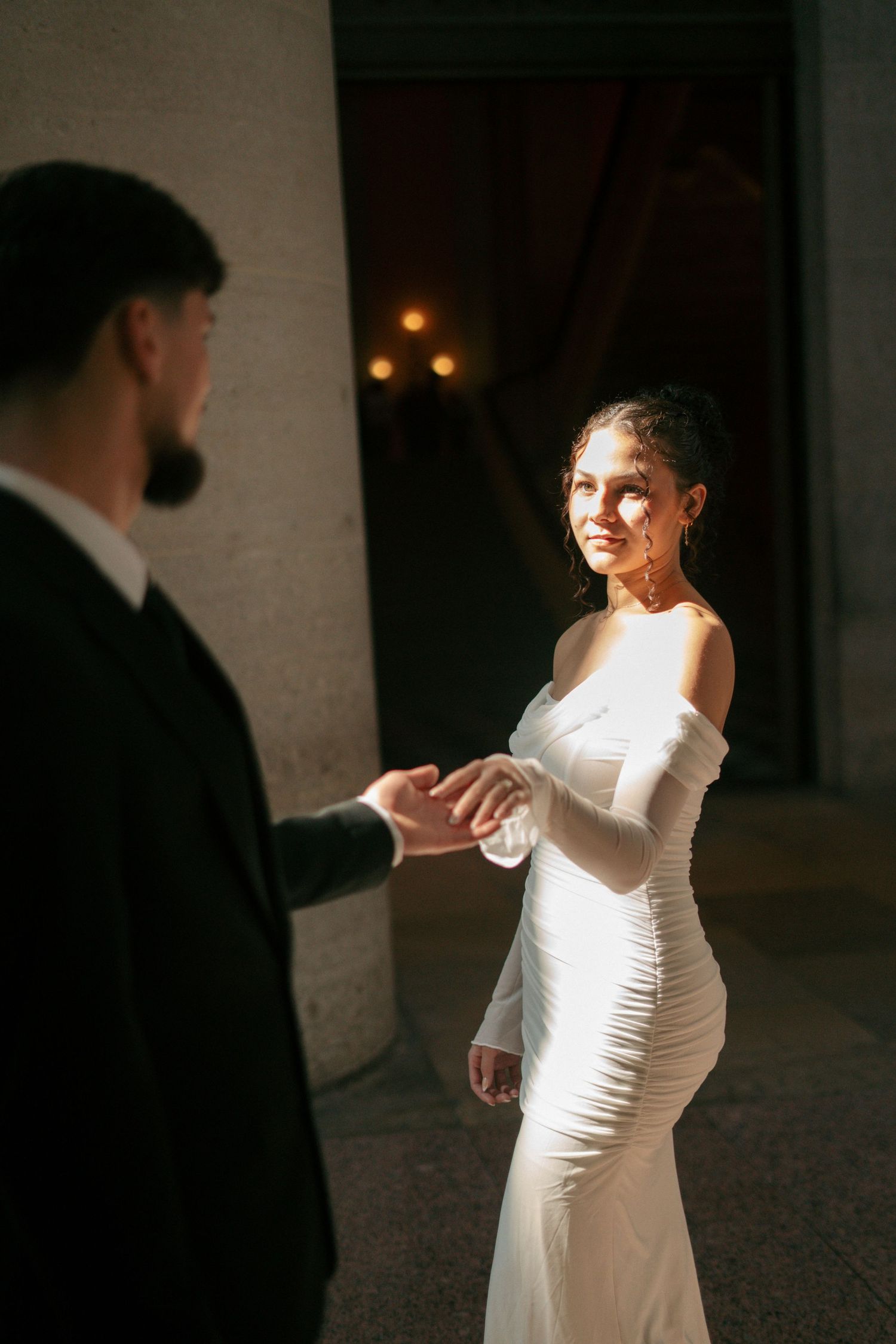 Bride in white off-shoulder gown holding grooms hand in dramatic light.