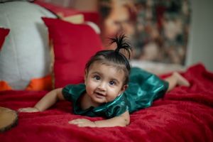 Baby in emerald green dress lying on red velvet couch with festive holiday decor.