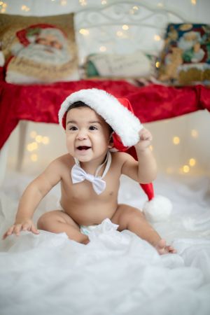 Baby in Santa hat and white bowtie sitting on fluffy white blanket with holiday lights backdrop.