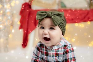 Baby wearing plaid shirt and green bow headband with joyful expression against holiday lights background.