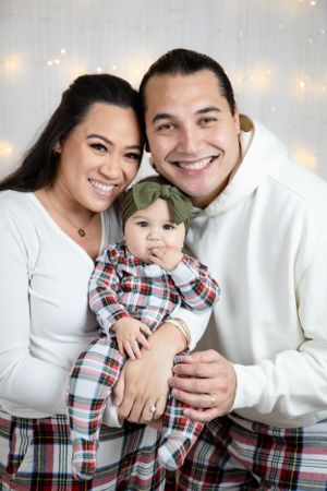 Happy family wearing coordinated holiday outfits pose together with string lights in background.