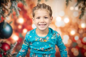 Child in blue holiday pajama top smiles brightly against Christmas backdrop with bokeh lights.
