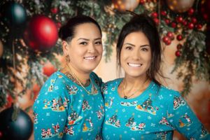 Two women in matching turquoise holiday sweaters smile in front of Christmas decorations.