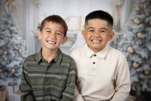 Two boys in casual attire share smiles in front of decorated Christmas tree backdrop.