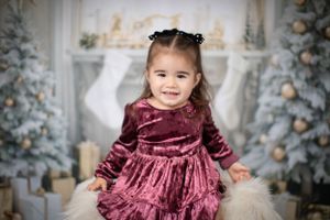 Small child in burgundy velvet dress poses by Christmas scene with trees and stockings.