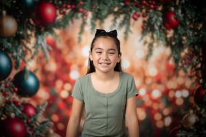 Young person in sage green top poses against festive backdrop with ornaments and bokeh lights.