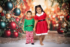 Children wearing festive Christmas outfits pose by decorated tree with ornaments and bokeh lights.