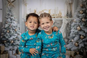 Two children in matching blue holiday pajamas pose in front of Christmas scene.